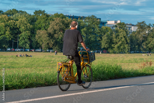 Yellow bike in the park: a man in headphones pedals a city bike with a basket on a sunny day. He enjoys the music, the fresh air and the green scenery of the park with trees and people.