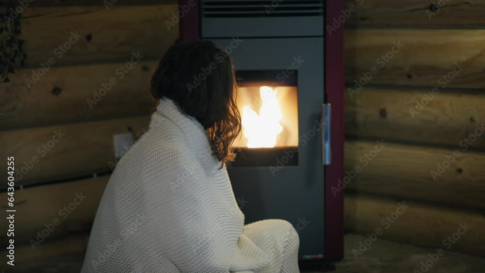 A young Caucasian woman is sitting by an electric pellet fireplace wrapped in a blanket. A young Caucasian woman is sitting by an electric pellet fireplace wrapped in a blanket.