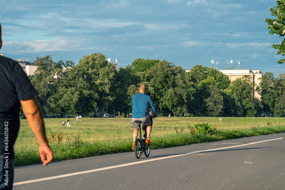 Obraz premium A man in a blue shirt and black shorts rides a black bicycle along a path surrounded by green trees and grass, past people walking in the park. In the background is a white building and a blue sky