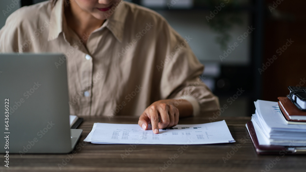 Female entrepreneur working to writing notes while reading document and finance chart on laptop Female entrepreneur working to writing notes while reading document and finance chart on laptop