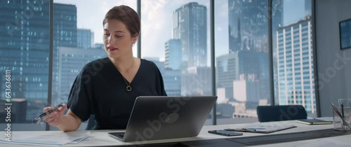 Portrait of Young Successful Caucasian Businesswoman Sitting at Desk Working on Laptop Computer in City Office. Female Manager Plan Investment Strategy for e-Commerce Project. Anamorphic Shot photo