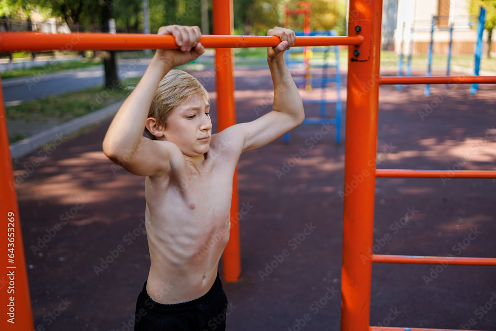 Obraz premium A teenager with athletic muscle development holds on to a bar. Street workout on a horizontal bar in the school park.
