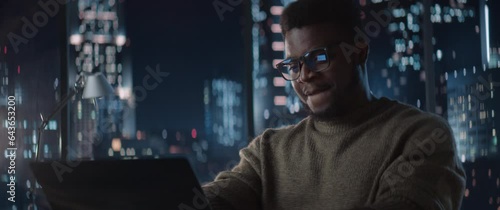 Successful Handsome Black Businessman Working on Laptop Computer in Big City Office Late at Night. Corporate Investment Manager Checking Financial Data from Project Management Report. Anamorphic Shot photo