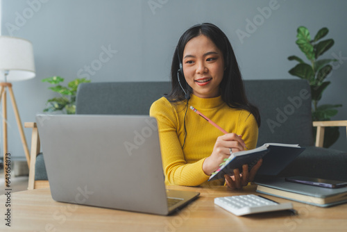 Attractive young Asian woman smiling happily at home studying internet video communication online using laptop computer with headset microphone typing data on keyboard to learn business education.