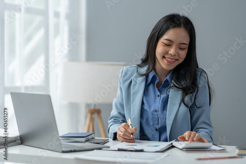 Young Asian accountant with a positive attitude using a calculator at her desk to calculate earnings. Check out the business growth report. accounting concepts, marketing