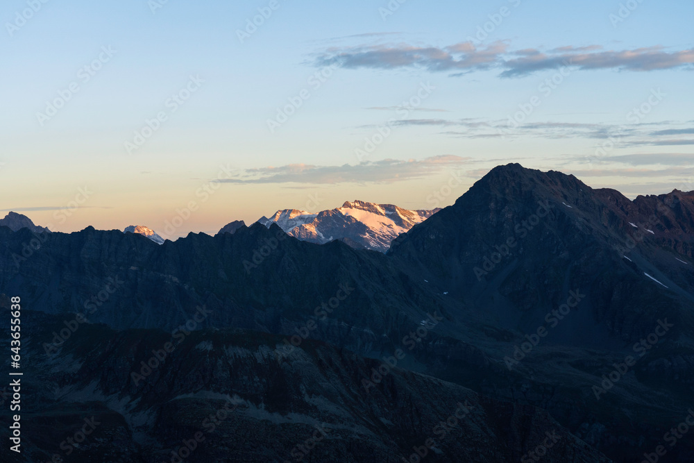 View of Testa del Rutor, Becca du Lac, Ruitor Massif from Gran Paradiso National Park glacier at sunrise. orange yellow colored peak in the background, foreground mountains and valley in the shadow