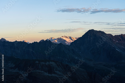 View of Testa del Rutor, Becca du Lac, Ruitor Massif from Gran Paradiso National Park glacier at sunrise. orange yellow colored peak in the background, foreground mountains and valley in the shadow