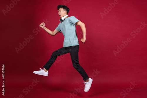 Carefree young guy in casual style with wireless headset jumping to the side on crimson color background,  full length, copy space © Queenmoonlite Studio