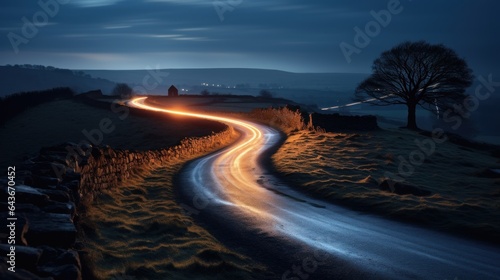 A curvy, winding country road with a path of light from leading headlights passes.