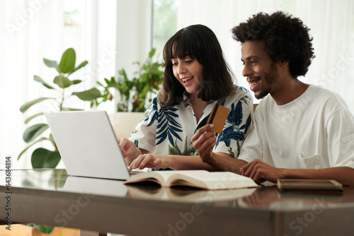 Happy young brunette woman choosing goods in online shop while sitting in front of laptop next ot African American boyfriend with credit card
