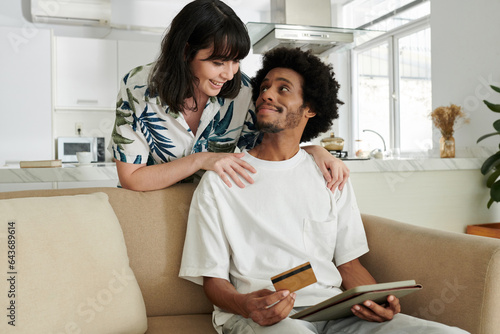 Young affectionate couple looking at one another while woman bending over her husband with tablet and credit card during online shopping
