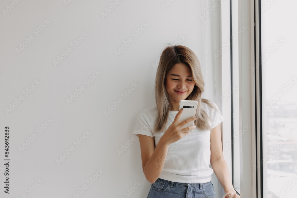 Asian woman smiling holding and looking at cellphone standing next to the window at apartment. Asian woman smiling holding and looking at cellphone standing next to the window at apartment.