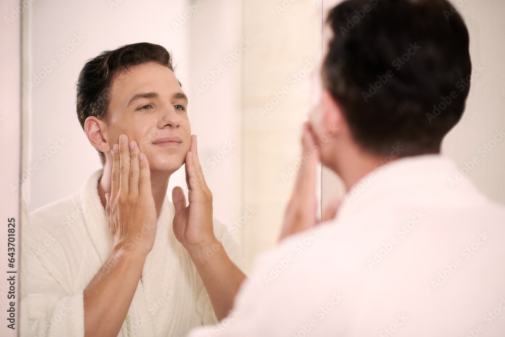 Young multiethnic man in white bathrobe touching his face while standing in front of mirror in bathroom and looking at his reflection Young multiethnic man in white bathrobe touching his face while standing in front of mirror in bathroom and looking at his reflection