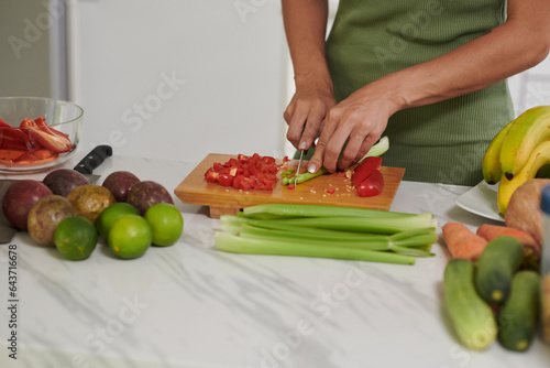 Hands of young woman cutting fresh celery and capsicum on wooden board while standing by kitchen table and cooking breakfast