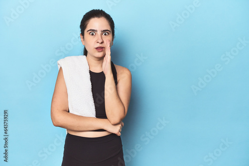 Sporty woman with towel on blue studio is saying a secret hot braking news and looking aside