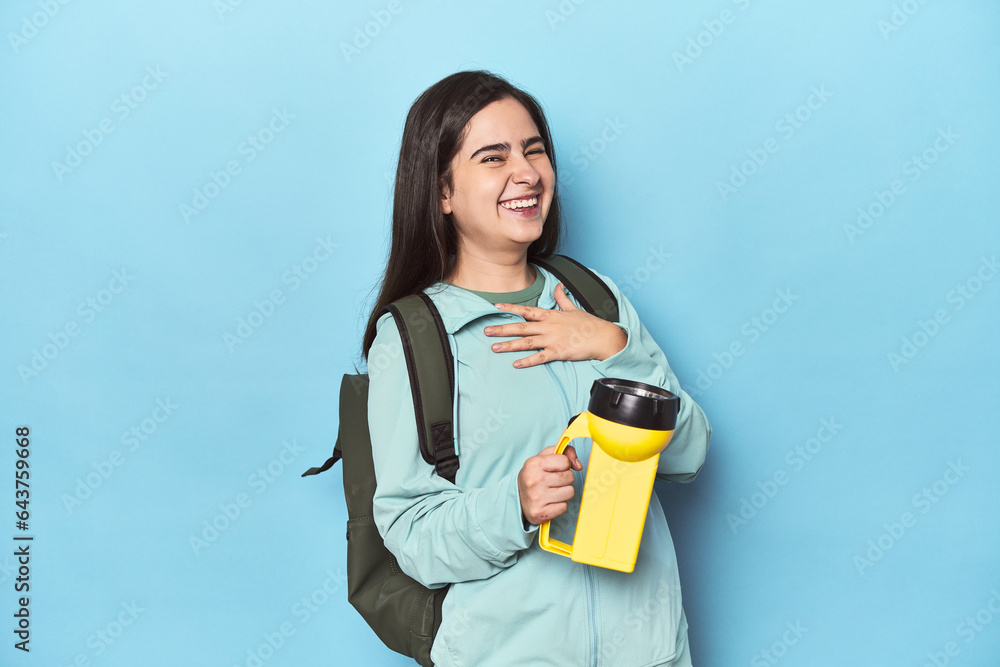 Adventurous woman with backpack on blue laughs out loudly keeping hand on chest. Adventurous woman with backpack on blue laughs out loudly keeping hand on chest.