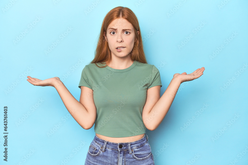 Redhead young woman on blue background confused and doubtful shrugging shoulders to hold a copy space. Redhead young woman on blue background confused and doubtful shrugging shoulders to hold a copy space.