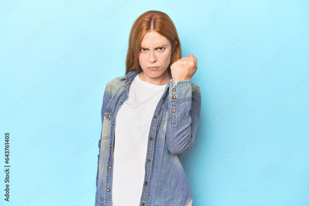 Redhead young woman on blue background showing fist to camera, aggressive facial expression. Redhead young woman on blue background showing fist to camera, aggressive facial expression.