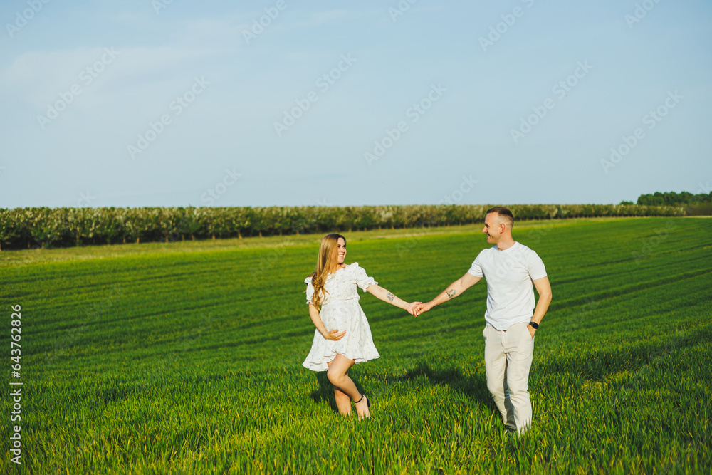 Pregnant couple hugging in a green spring field. Expectant parents in the park of white flowering trees. A romantic couple expecting a baby. A walk through a green field Pregnant couple hugging in a green spring field. Expectant parents in the park of white flowering trees. A romantic couple expecting a baby. A walk through a green field