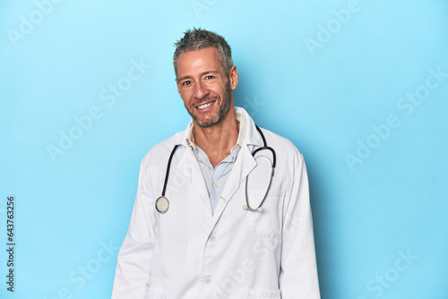 Caucasian middle-aged doctor on blue background happy, smiling and cheerful.