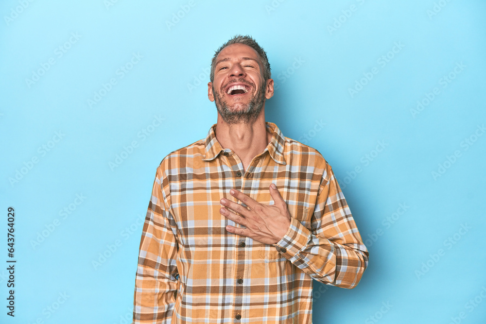 Middle-aged caucasian man on blue backdrop laughs out loudly keeping hand on chest. Middle-aged caucasian man on blue backdrop laughs out loudly keeping hand on chest.