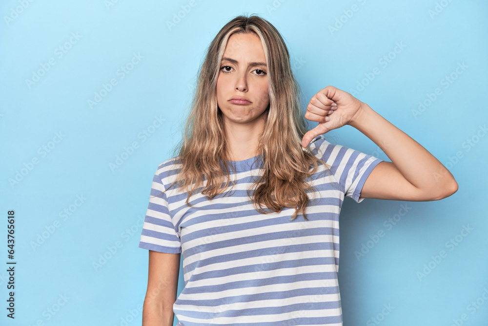 Blonde young caucasian woman in blue studio showing a dislike gesture, thumbs down. Disagreement concept. Blonde young caucasian woman in blue studio showing a dislike gesture, thumbs down. Disagreement concept.