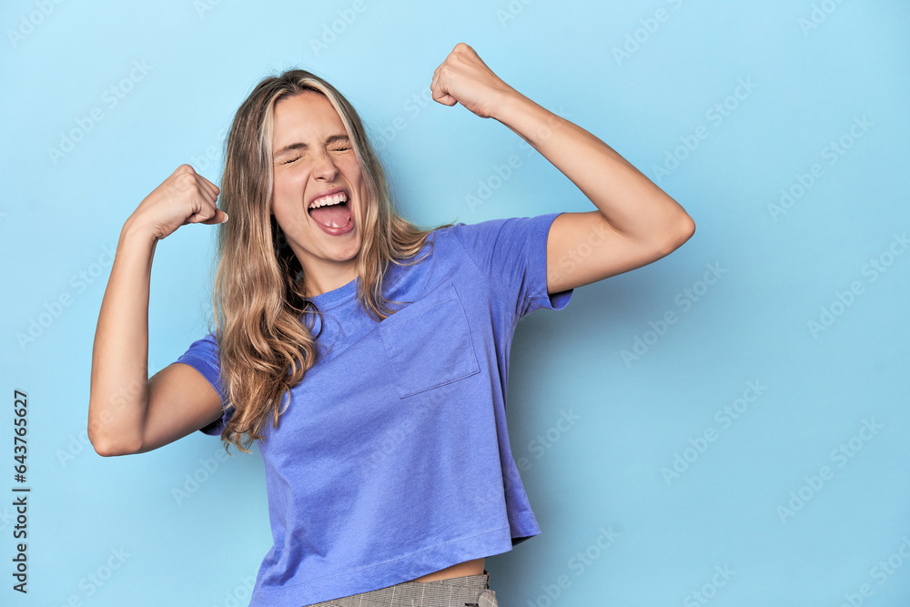 Fototapeta premium Blonde young caucasian woman in blue studio raising fist after a victory, winner concept.