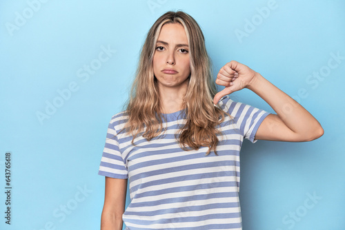 Blonde young caucasian woman in blue studio showing a dislike gesture, thumbs down. Disagreement concept.