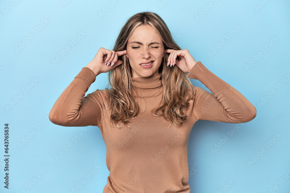 Blonde young caucasian woman in blue studio covering ears with fingers, stressed and desperate by a loudly ambient. Blonde young caucasian woman in blue studio covering ears with fingers, stressed and desperate by a loudly ambient.