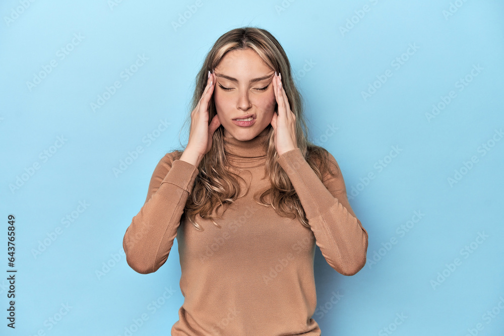 Blonde young caucasian woman in blue studio touching temples and having headache. Blonde young caucasian woman in blue studio touching temples and having headache.