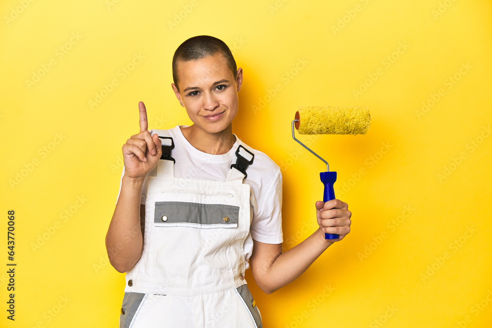 Painter woman, shaved head, with yellow roller in studio showing number one with finger. Painter woman, shaved head, with yellow roller in studio showing number one with finger.