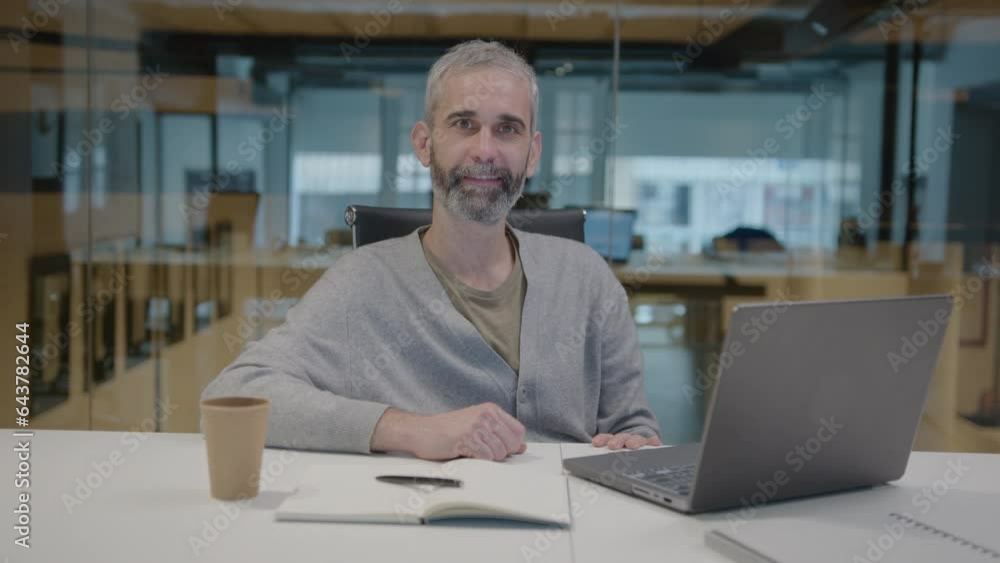 Senior businessman with gray hair sitting at office desk with laptop and copybook, posing on camera and smiling. Medium shot, video portrait Senior businessman with gray hair sitting at office desk with laptop and copybook, posing on camera and smiling. Medium shot, video portrait