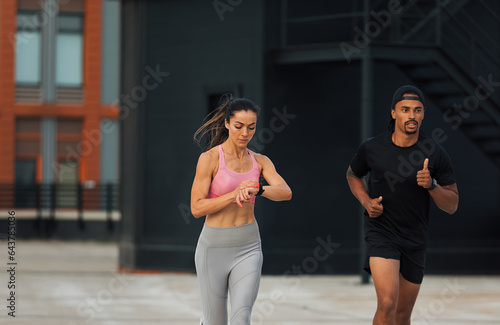 Two athletes sprinting on a rooftop. Young woman checking fitness tracker while running.