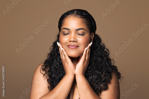 Perfect skin. Beautiful brazilian plus size woman touching her face with pleased face expression, brown background