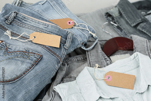 Stack of blue jeans, shirts and empty tag. Second hand clothing shop. Circular fashion, eco friendly sustainable shopping, donation, thrift stores concept. Top view with tag mockup photo