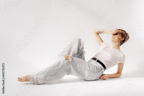 Side view of young female lying on floor with raised leg and back while using support of hands and looking away wearing a simple white t-shirt and light blue jeans against white background