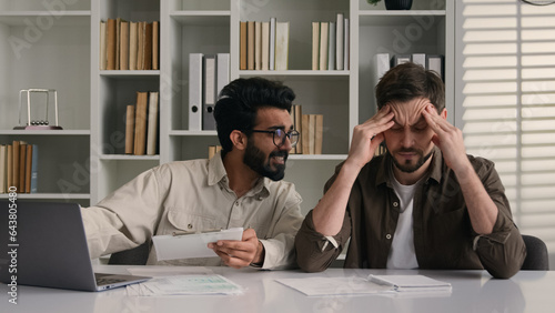 Overworked tired sick man suffer headache ignoring obsessive annoying colleague disturb talk smile discuss project with laptop two multiethnic businessmen diverse men solve teamwork problem in office