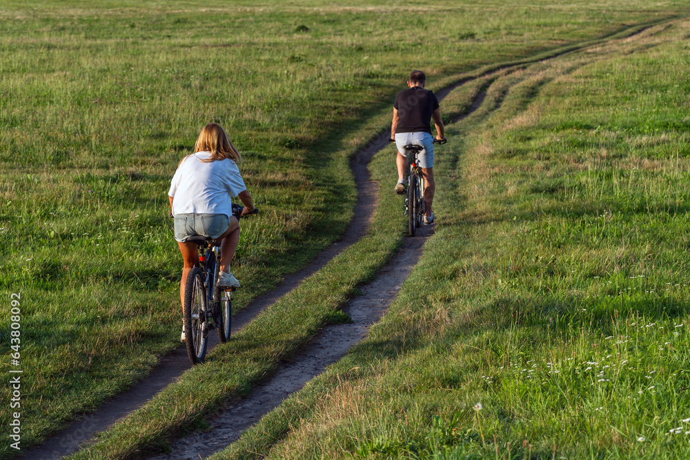 Two cyclists, a girl and a boy, are enjoying a ride on a rural dirt road. They wear casual clothes and are surrounded by green fields and trees. The picture captures a moment of leisure and relaxation