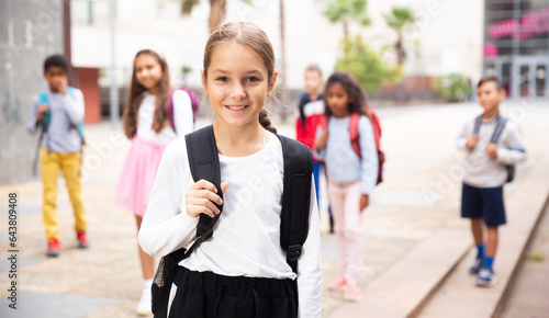 Portrait of positive schoolgirl standing near school, children on background