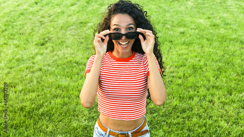Lovely Mixed Race curly woman looking directly at the camera laughing, facial expression happiness. Cute girl took off her glasses and laughs fooling around in the park in nature grass background