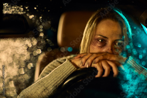 A young Caucasian woman, marked by the harrowing evidence of domestic abuse with distinct bruises and darkened eyes, sits in her car, leaning heavily on the steering wheel. photo