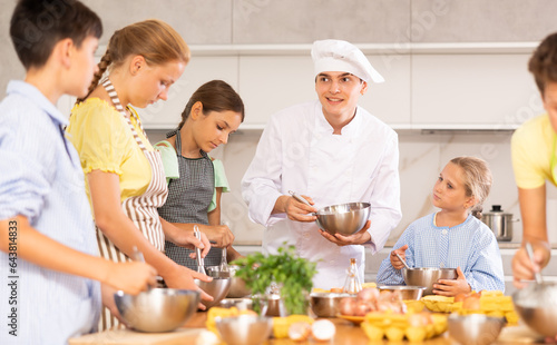 Friendly positive young guy  qualified chef  running culinary courses for group of tween children  sharing professional secrets of cooking ..