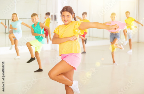 Portrait of cheerful preteen hispanic girl learning energetic dance moves with group of children in choreography class .