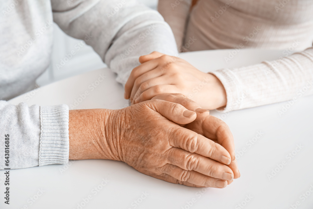 Fototapeta premium Young woman with her grandmother holding hands at table in kitchen, closeup