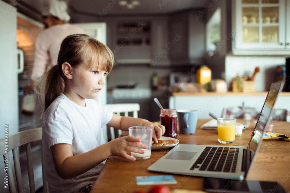 Young daughter having breakfast with her mother while using the laptop in the kitchen