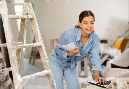 Positive female caucsaian architect holding documents, using laptop during repair works in house. photo