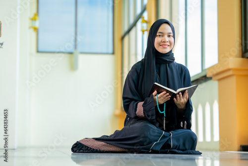 The image of an Asian Muslim woman in the Islamic religion in hijab in black color. reading the Quran and having a happy smiling face Staying in a beautiful mosque out of respect for God.