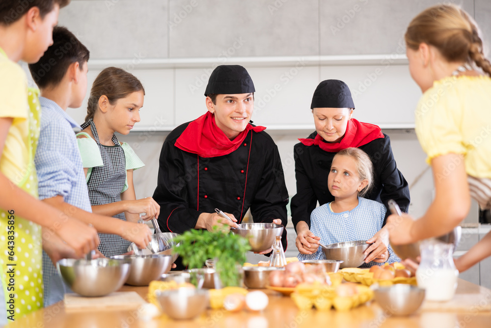 Cheerful enthusiastic woman and young man, skilled chefs wearing black jackets and caps, conducting culinary masterclass for teenage children, sharing secrets of cooking