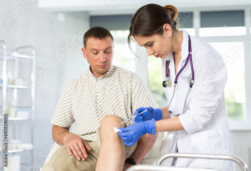 Young female doctor performs intra-articular injection treatment of knee bursitis to middle-aged man patient