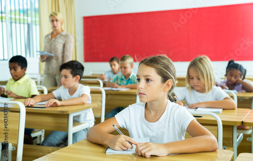 Portrait of positive small female pupil sitting at desk studying in classroom